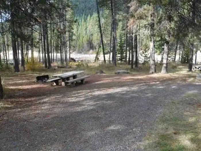 Flat gravel parking area next to picnic table and fire ring in pine forest with lake in the background.